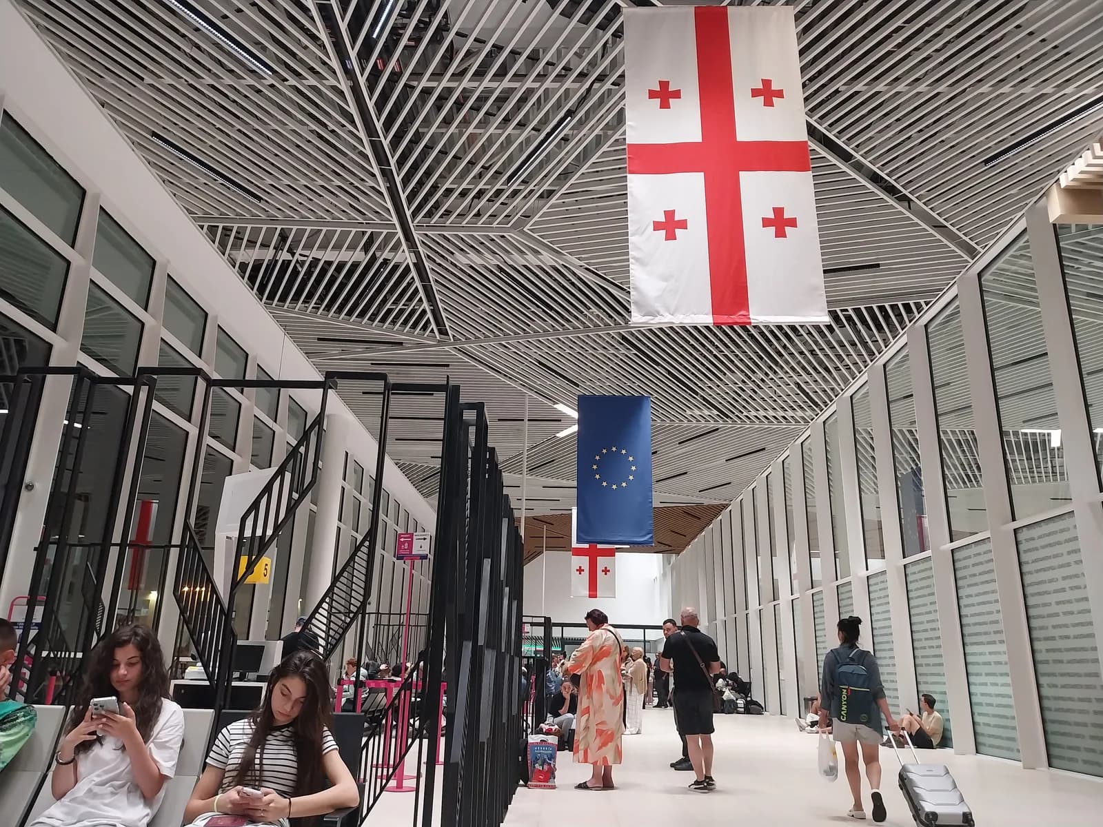 The departure concourse at Kutaisi International Airport, seating and gates along one glazed corridor beneath hanging Georgian and EU flags